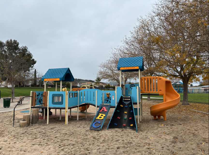 Playground featuring slides, ramps, and climbing elements with surrounding trees and pathways.
