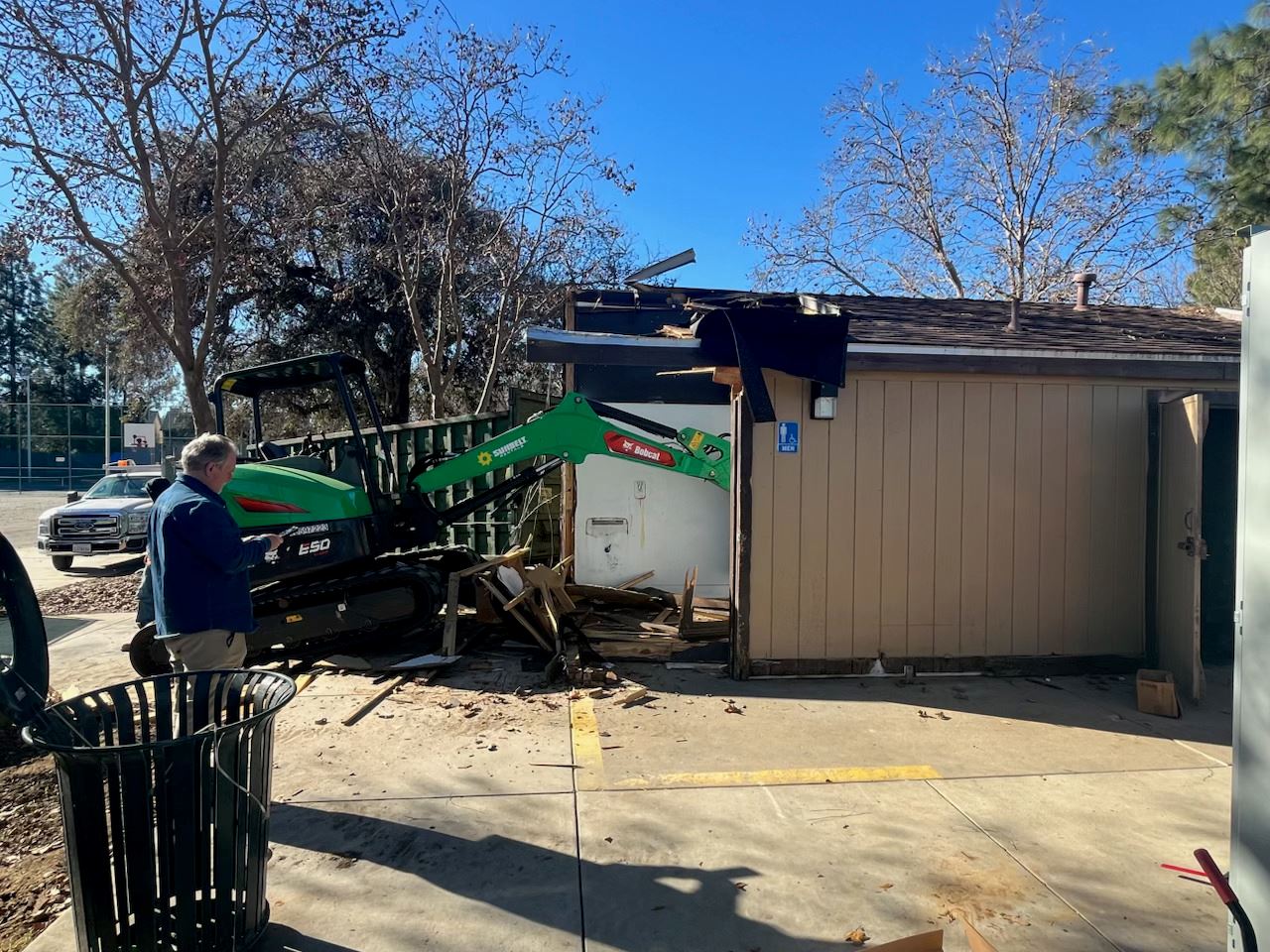 Excavator demolishing a small park restroom building while a worker observes nearby.